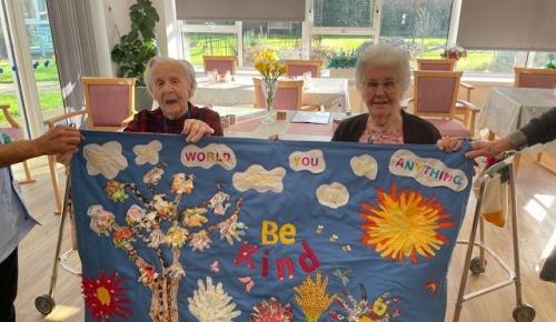 Norfolk care home residents Mary and Norma hold up a piece of textile art created in collaboration with local children. A rainbow coloured tree and flower scene created from handprints on a blue fabric background.