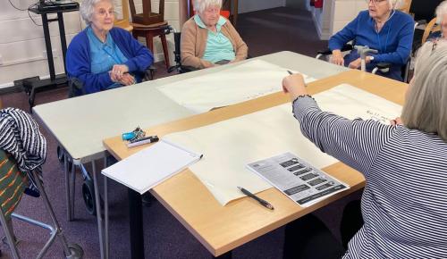 A group of Norwich care home residents discuss what makes them feel safe living in Corton House with staff members. There are papers on the table and everyone looks thoughful.