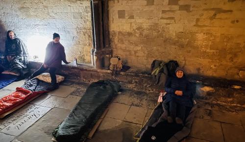 Corton House staff members sit in sleeping bags in Norwich cathderal cloister. They are wrapped up warm and smiling.