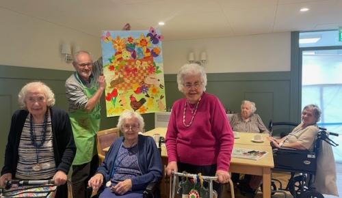 Residents at Norwich care home Corton House show off a painted collage they have created for Easter. They are standing and sitting in a panelled dining room with green walls and parquet flooring, holding the art and smiling.