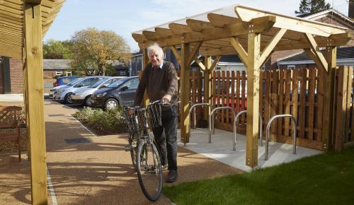 A tenant at Brakendon Close Independent Living Scheme in Norwich stands in the garden and car park area of the sheltered housing complex, smiling with his bike next to a purpose built bike shed.