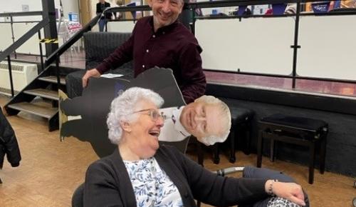 The manager of Norwich care home Corton House smiles and jokes with resident Norma, he is holding a life size cut out of King Charles. Both are laughing.